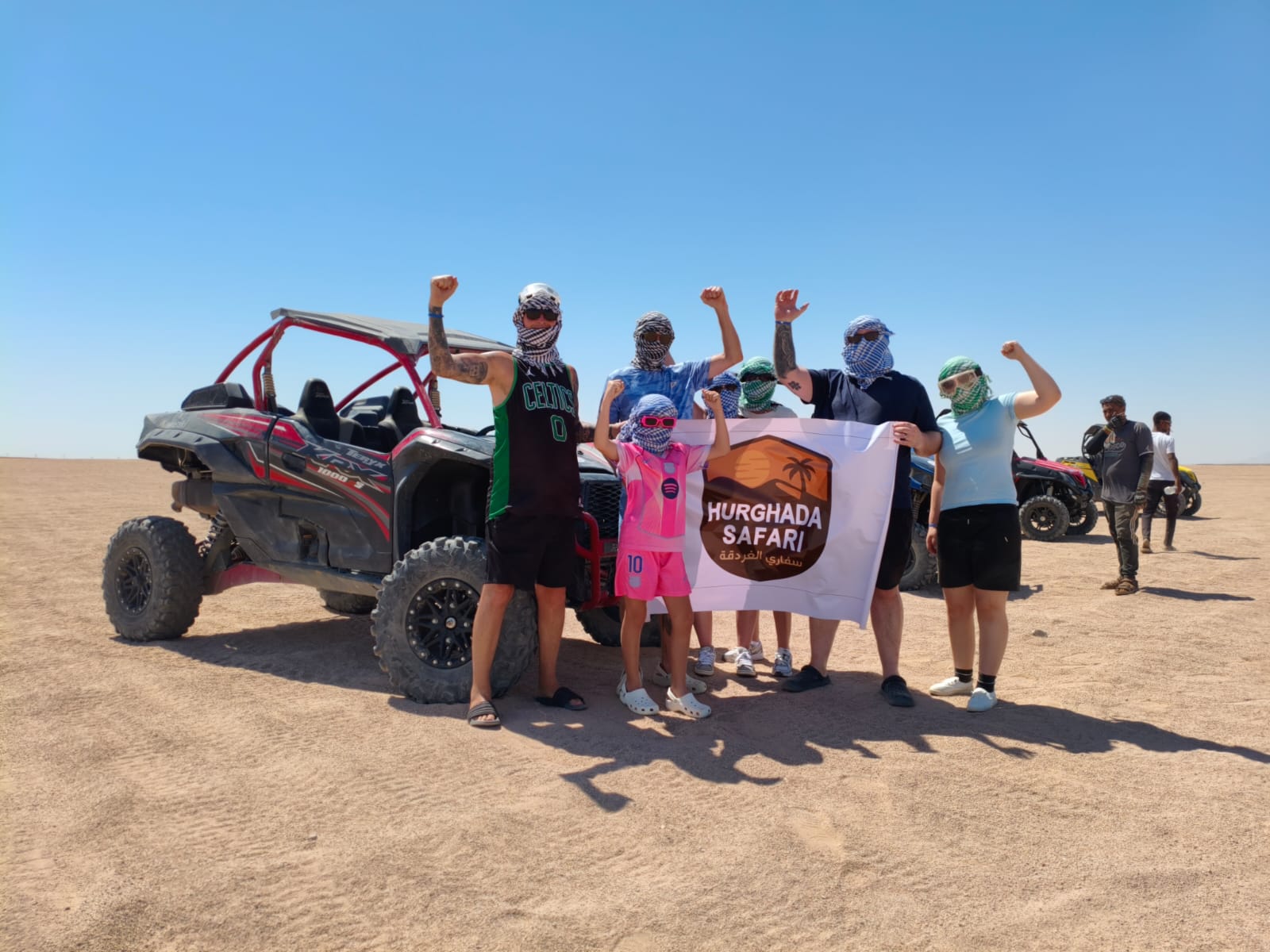 Group of Hurghada Safari guests celebrating beside a buggy holding the Hurghada Safari flag in the desert